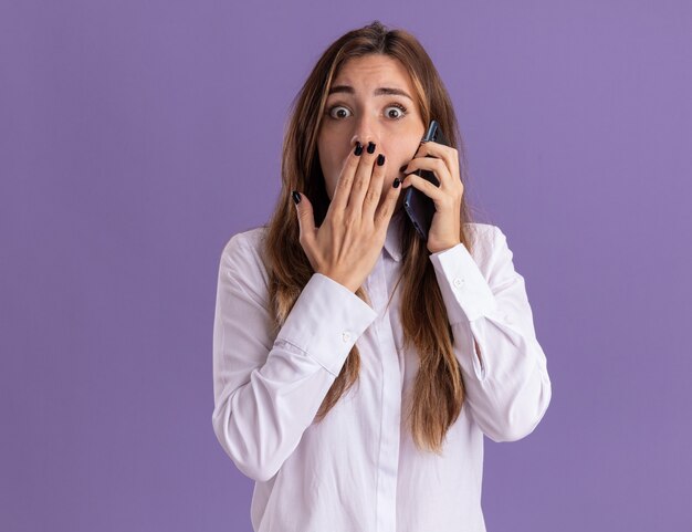 불안한 젊은 백인 소녀는 입에 손을 대고 복사 공간이 있는 보라색 벽에 격리된 전화로 이야기합니다