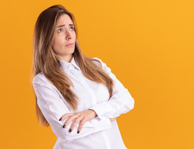 Anxious young pretty caucasian girl stands with crossed arms looking at side isolated on orange wall with copy space