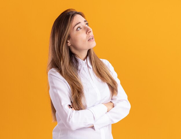 Anxious young pretty caucasian girl stands with crossed arms looking up isolated on orange wall with copy space