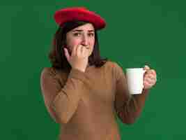 Foto gratuita ansiosa giovane ragazza abbastanza caucasica con cappello berretto morde le unghie e tiene la tazza sul verde