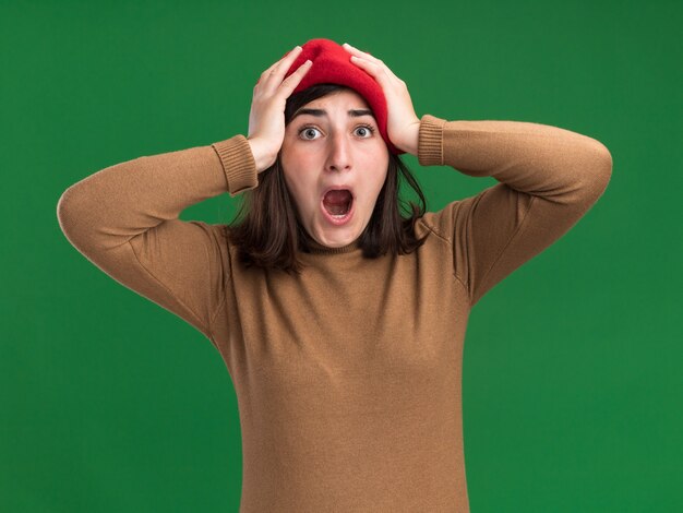 Anxious young pretty caucasian girl with beret hat puts hands on head on green