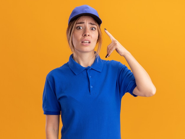 Anxious young pretty delivery girl in uniform points up on orange