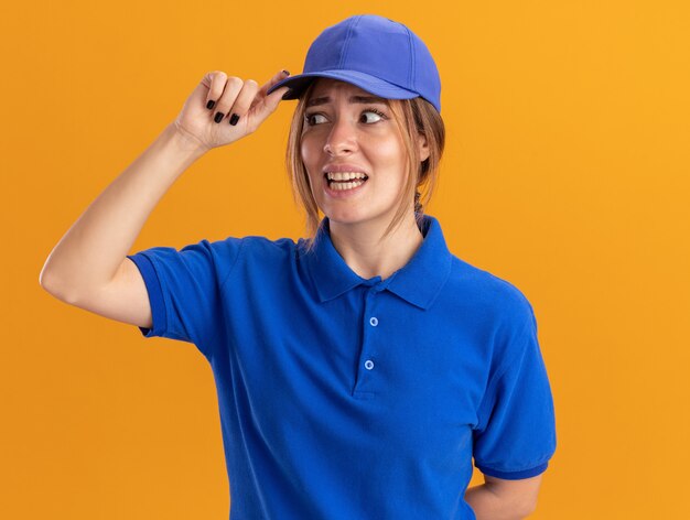 Anxious young pretty delivery girl in uniform puts hand on cap and looks at side on orange