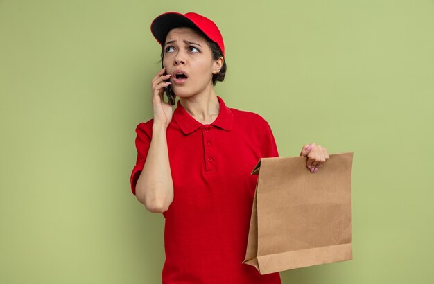 Тревожная молодая красивая женщина-доставщик разговаривает по телефону и держит бумажный пакет с едой, глядя в сторону