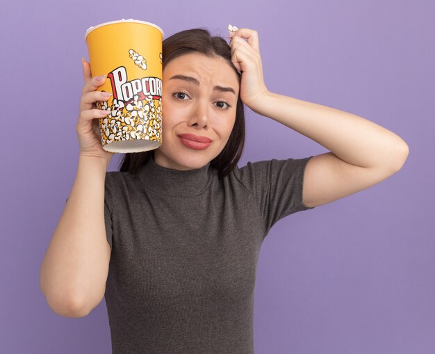 Обеспокоенная молодая красивая женщина, держащая ведро с попкорном и кусок попкорна, касается головы ведром попкорна и рукой, смотрящей вперед, изолированной на фиолетовой стене