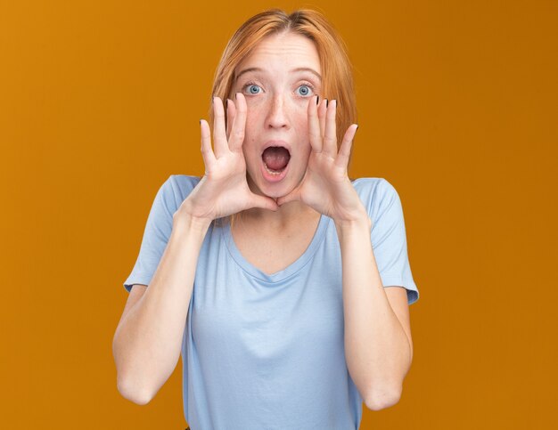 Anxious young redhead ginger girl with freckles holding hands close to mouth on orange