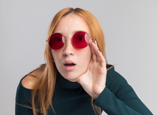 Обеспокоенная молодая рыжая рыжая девушка с веснушками в солнцезащитных очках кладет руку на лицо, изолированное на белой стене с копией пространства