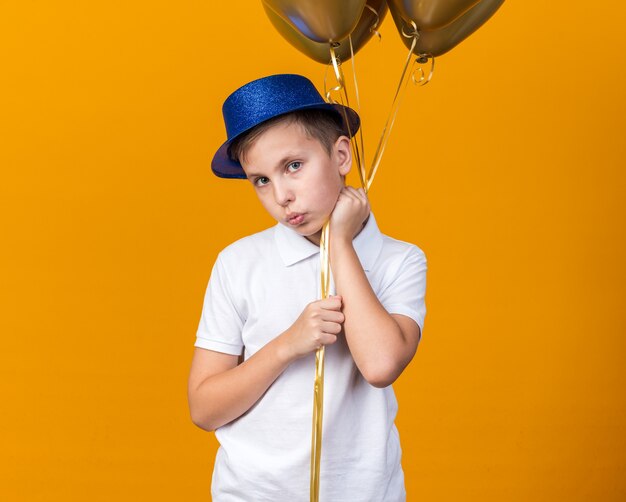 Ansioso giovane ragazzo slavo con cappello da festa blu che tiene palloncini di elio isolati sulla parete arancione con spazio di copia