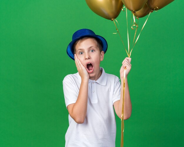 anxious young slavic boy with blue party hat putting hand on face and holding helium balloons isolated on green wall with copy space