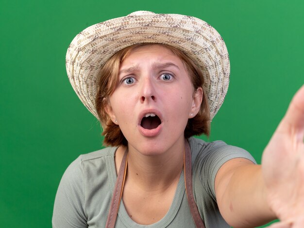 Anxious young slavic female gardener wearing gardening hat looking at camera pretending to hold camera on green