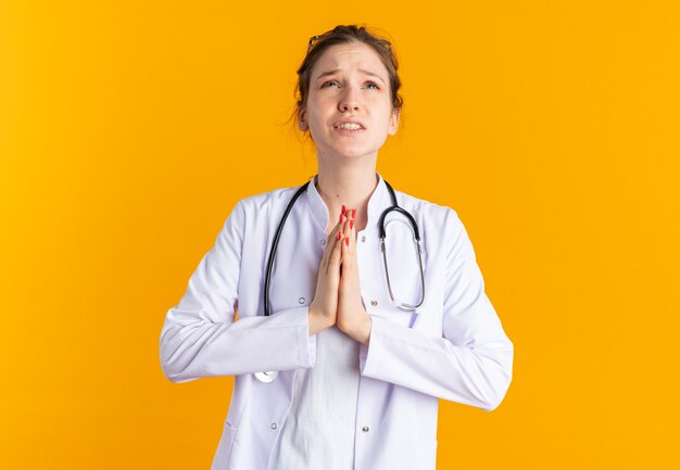 Anxious young slavic girl in doctor uniform with stethoscope praying looking up isolated on orange wall with copy space