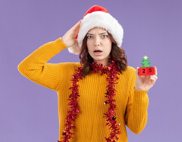 산타 모자와 목에 갈 랜드와 함께 불안한 젊은 슬라브 소녀는 머리에 손을 넣고 복사 공간이 보라색 배경에 고립 된 크리스마스 트리 장식을 보유하고 있습니다.