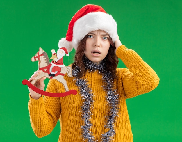 산타 모자와 목에 갈 랜드와 함께 불안한 젊은 슬라브 소녀는 머리에 손을 넣고 복사 공간이 녹색 벽에 고립 된 흔들 목마 장식에 산타를 보유하고 있습니다.