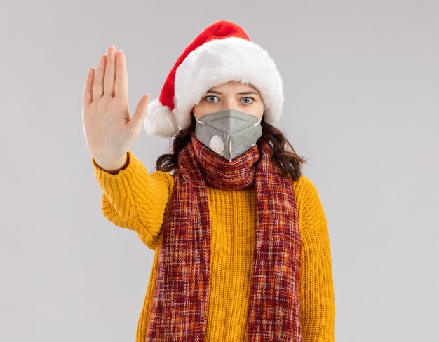 Anxious young slavic girl with santa hat and with scarf around neck wearing medical mask gesturing stop hand sign isolated on white wall with copy space