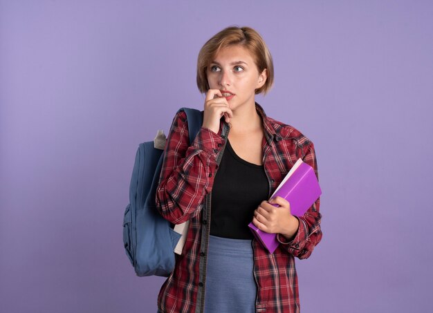 Anxious young slavic student girl wearing backpack bites nail holds book and notebook 