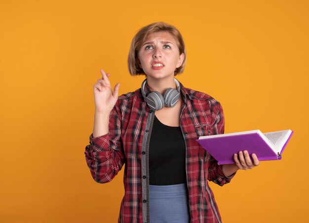 Anxious young slavic student girl with headphones wearing backpack crosses fingers and holds book 