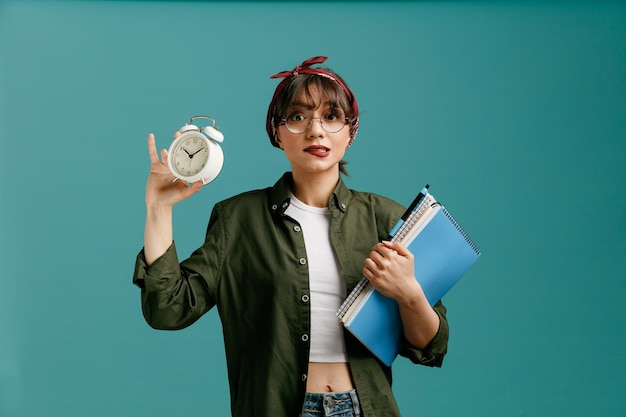 Anxious young student girl wearing bandana glasses holding large note pads with pen looking at camera biting lip showing alarm clock isolated on blue background