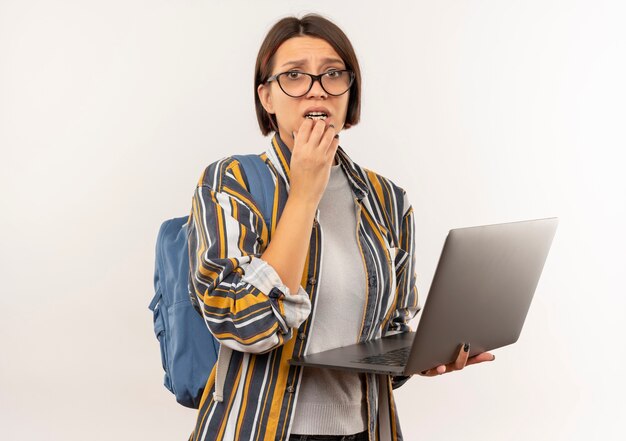 Anxious young student girl wearing glasses and back bag holding laptop putting hand on chin isolated on white  with copy space