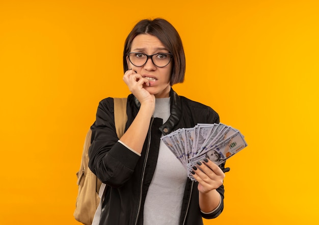 Anxious young student girl wearing glasses and back bag holding money putting hand on chin isolated on orange  with copy space