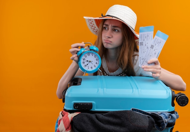 Взволнованная молодая путешественница в шляпе держит билеты на самолет и будильник с чемоданом на изолированном оранжевом пространстве с копией пространства