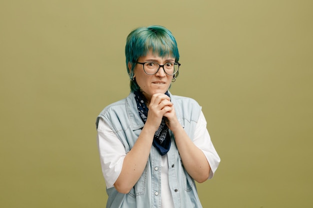 Ansiosa giovane donna che indossa occhiali bandana sul collo tenendo le mani insieme sul mento guardando la fotocamera isolata su sfondo verde oliva