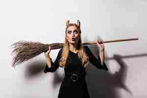 Free photo appealing black witch holding broom on white wall.  attractive blonde woman enjoying halloween party.