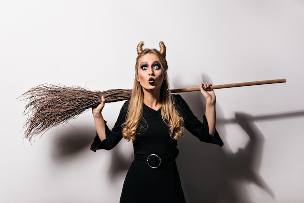 Appealing black witch holding broom on white wall.  attractive blonde woman enjoying halloween party.