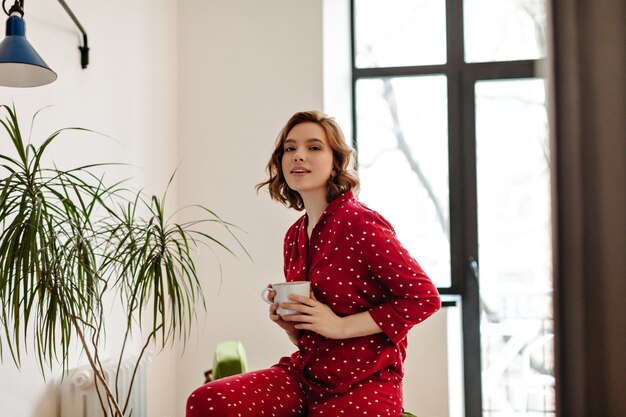 Appealing european woman in pajama holding cup of coffee and looking at camera. Indoor shot of young woman posing in morning at home.