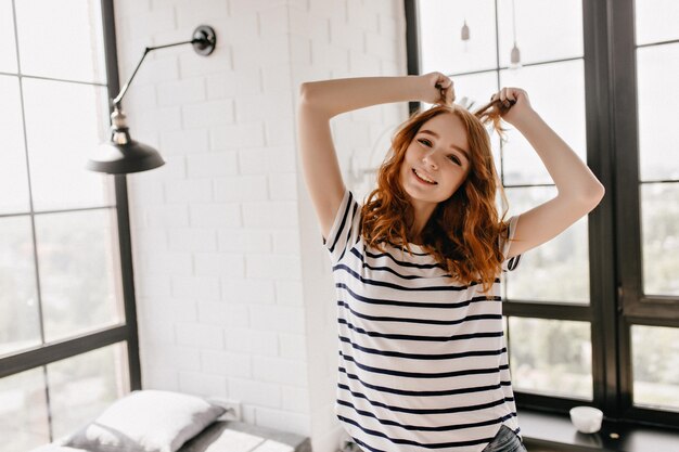 Appealing girl with wavy hair posing in her cozy room. Graceful curly ginger lady dancing at home.