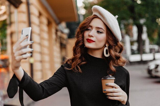 Привлекательная длинноволосая девушка использует телефон для селфи на улице. Удивительная женщина имбиря, наслаждаясь кофе на открытом воздухе.