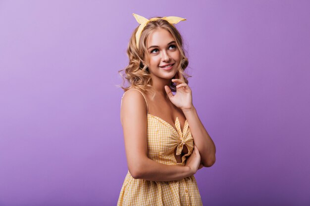 Appealing tanned girl posing on purple with dreamy face expression. Indoor portrait of winsome blonde woman in yellow dress.