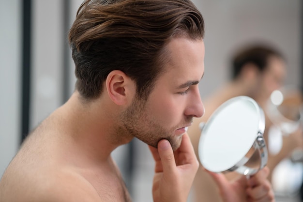 Free photo appearance. young man examining his face while watching into the round mirror