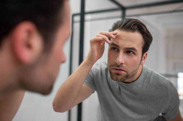 Appearance. A young man in grey tshirt looking at his reflection in the mirror