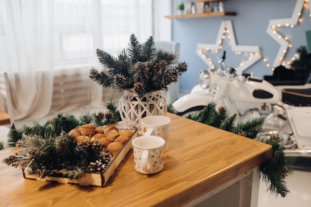 Appetizing cupcakes and mug on table serving Christmas decoration at cosiness room interior. Cozy holiday studio decorated by light and spruce white decorative motorbike at background