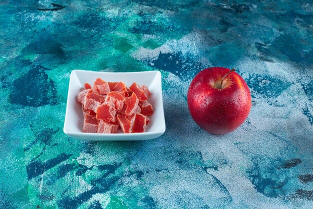 Яблоко и красный мармелад в миске на синем столе.
