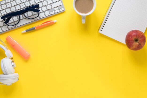 An apple; tea cup; keyboard; eyeglasses; pen; spiral notepad and white headphone on yellow backdrop