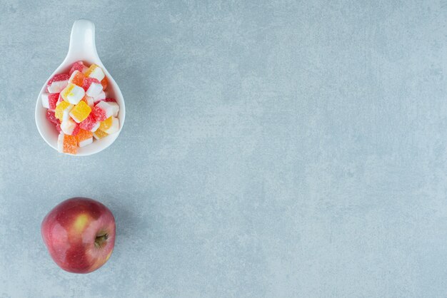 An apple and a tiny bowl of candy on marble.