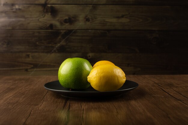 An apple and two lemons in a dark plate.