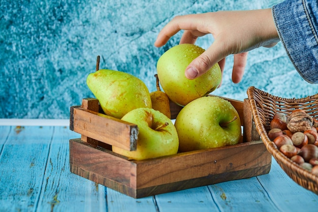 Apples and pear in wooden basket and bowl of hazelnuts on blue surface.