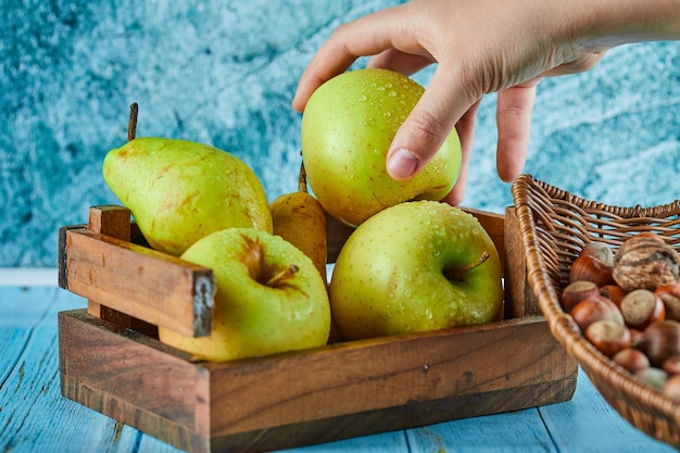 Яблоки и груши в деревянной корзине и шаре фундуков на синем столе.