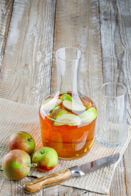 Apples with juice, knife, glass on wooden and kitchen towel, high angle view.