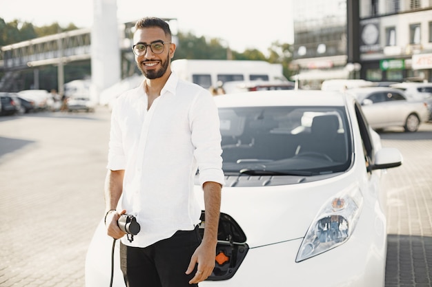 Arab man holding electric car plug. Smiling at camera, standing in the city near electro car.