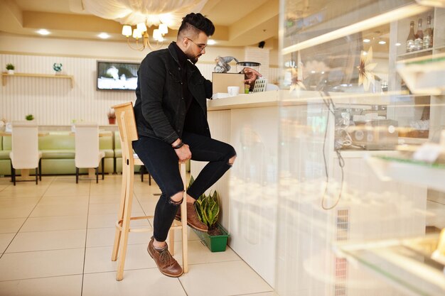 Arab man wear on black jeans jacket and eyeglasses in cafe drink coffee at bar with book Stylish and fashionable arabian model guy
