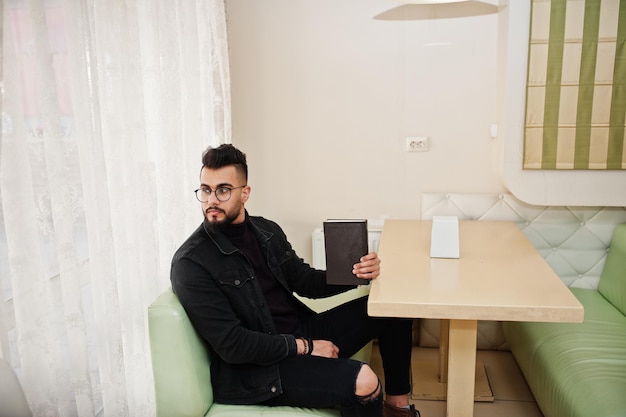 Arab man wear on black jeans jacket and eyeglasses sitting in cafe read book Stylish and fashionable arabian model guy