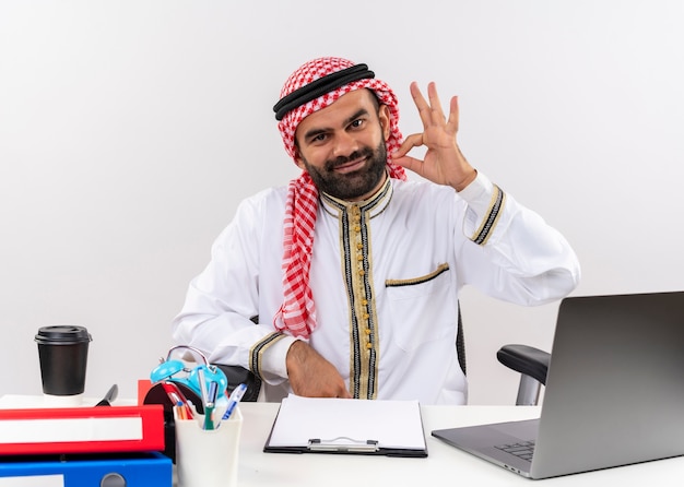 Бесплатное фото Арабский бизнесмен в традиционной одежде, сидя за столом с портативным компьютером, счастлив и позитивен, делает хорошо знаком, работая в офисе