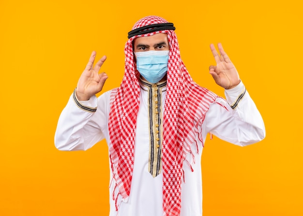 오렌지 벽 위에 서있는 손가락으로 명상 제스처를 만드는 자신감 표현으로 전통적인 착용과 얼굴 보호 마스크 아랍어 사업가