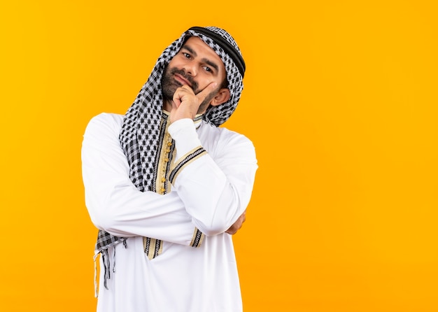 オレンジ色の壁の上に立って幸せで前向きな笑顔の伝統的な服を着たアラビアのビジネスマン
