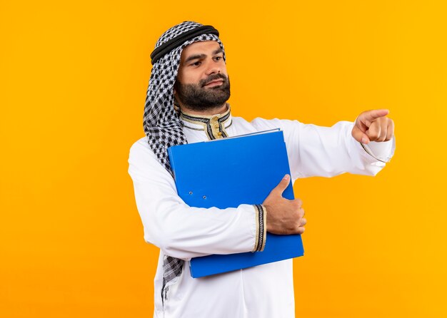 Arabic businessman in traditional wear holding blue folder looking aside pointing with finger to the side standing over orange wall