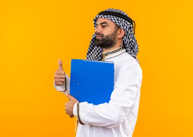 Arabic businessman in traditional wear holding blue folder showing thumbs up standing sideways over orange wall