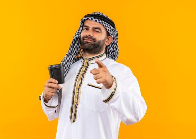 オレンジ色の壁の上に立って指をまばたきと笑顔で指しているコーヒーカップを保持している伝統的な服を着たアラビアのビジネスマン